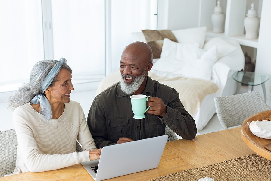 Front view of diverse senior couple using laptop on table while man holds a cup in beach house.